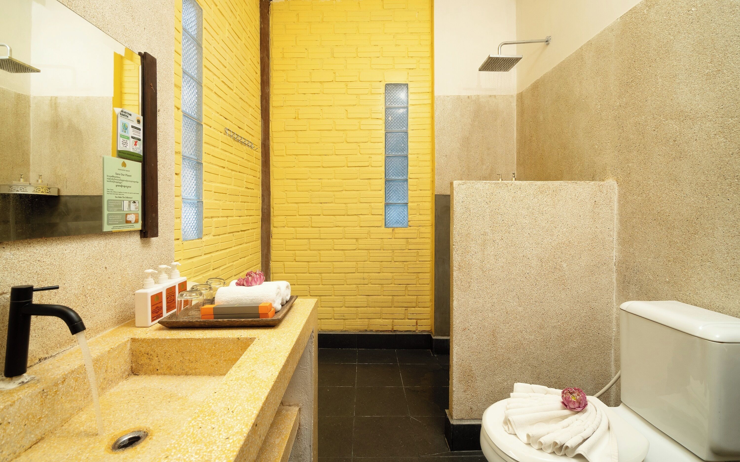 Un baño con pared de ladrillo amarillo, un lavamanos con artículos de aseo y un inodoro con toallas.