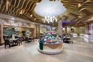 A spacious hotel lobby with a central reception desk, multiple dining areas, and a modern ceiling design.