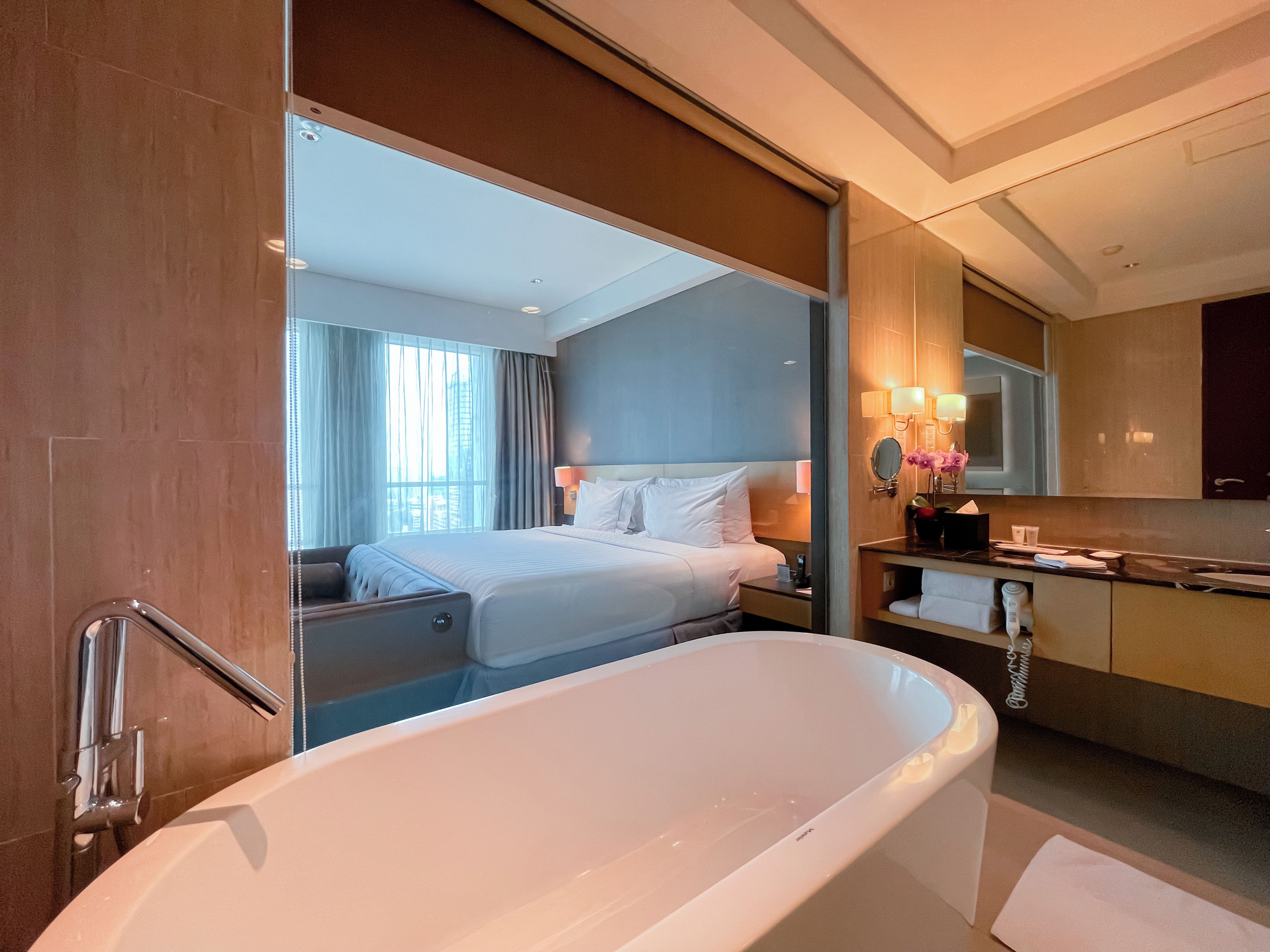 Una habitación de hotel moderna con una bañera grande, una cama bien hecha, un tocador con espejo y vistas a la ciudad desde la ventana.
