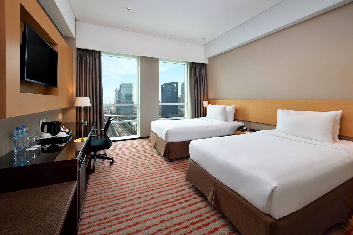 A hotel room with two beds, a desk with a flat-screen TV, a coffee maker, and a view of the cityscape.