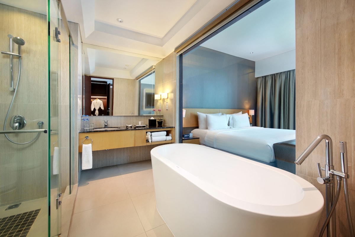 A modern hotel room with a glass shower enclosure, a large bathtub, a wooden vanity with a mirror, and a bed with white linens.