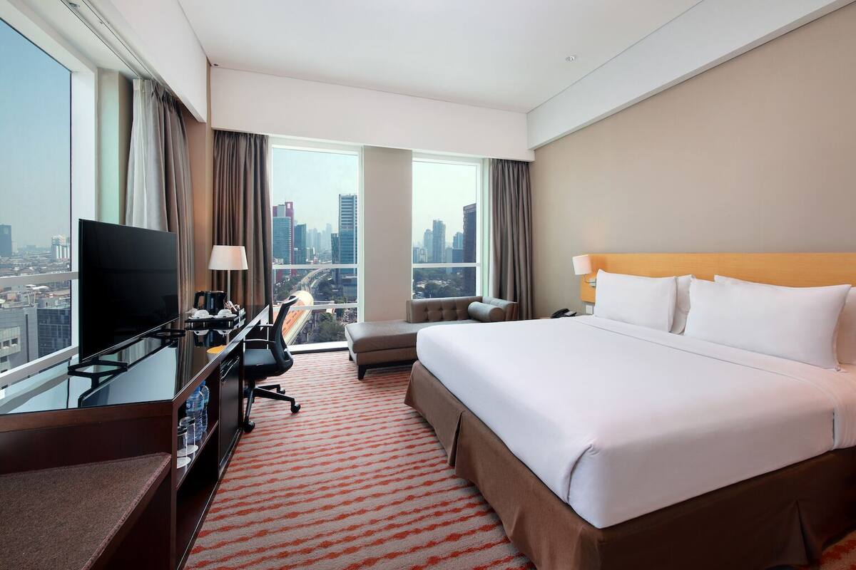 A hotel room with a large bed, a desk with a TV, a chair, and a view of the cityscape.