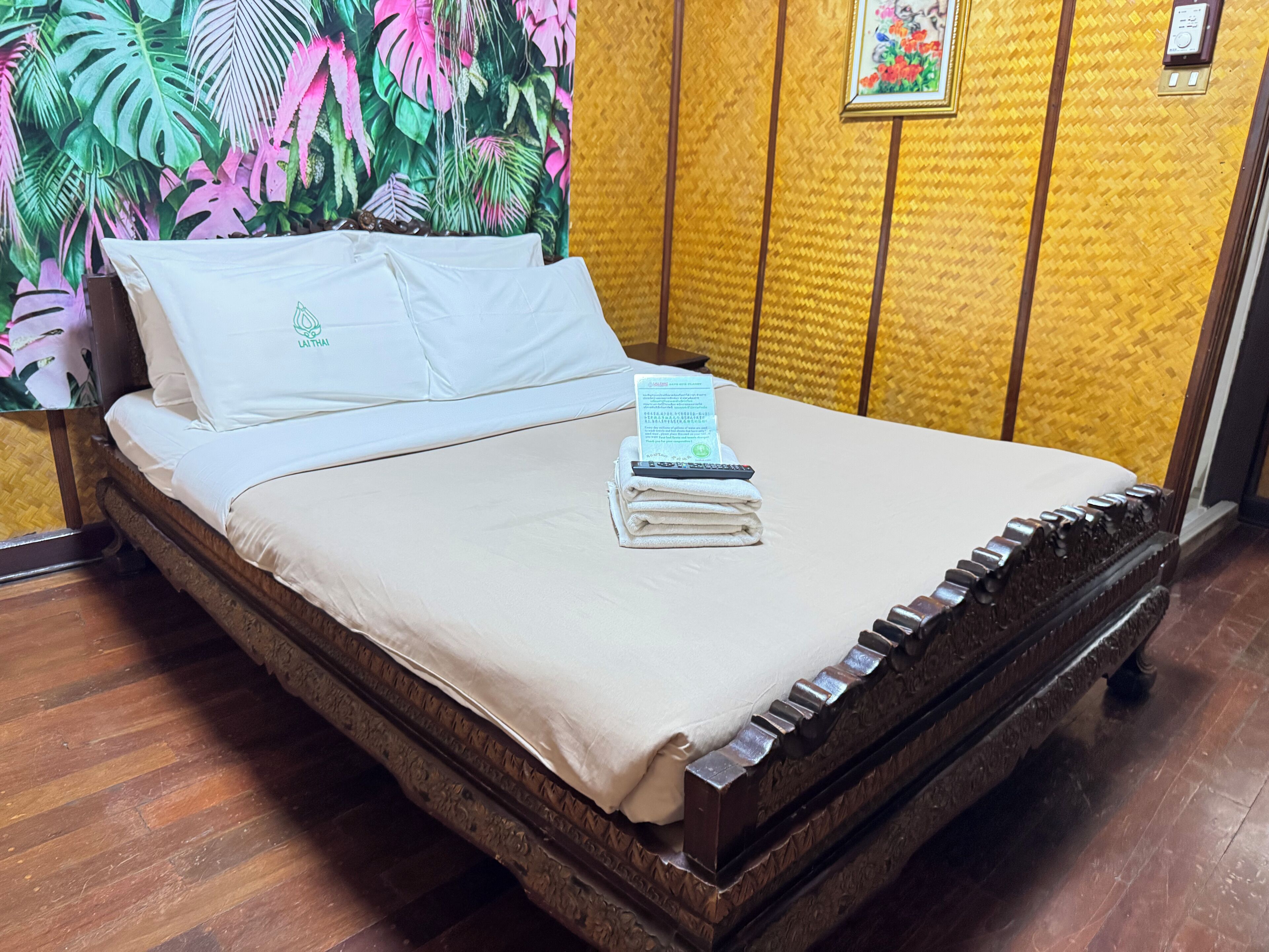 A bed with a white bedspread, pillows, and a folded towel. There is a remote control and a book on the bed. The room has wooden flooring and a yellow textured wall.
