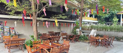 An outdoor seating area with wooden tables and chairs, surrounded by trees and decorated with colorful paper lanterns.