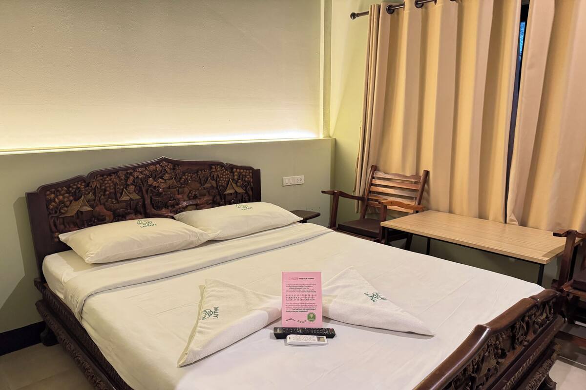 Een net opgemaakt bed met kussens, een nachtkastje, een stoel en een raam met gordijnen.