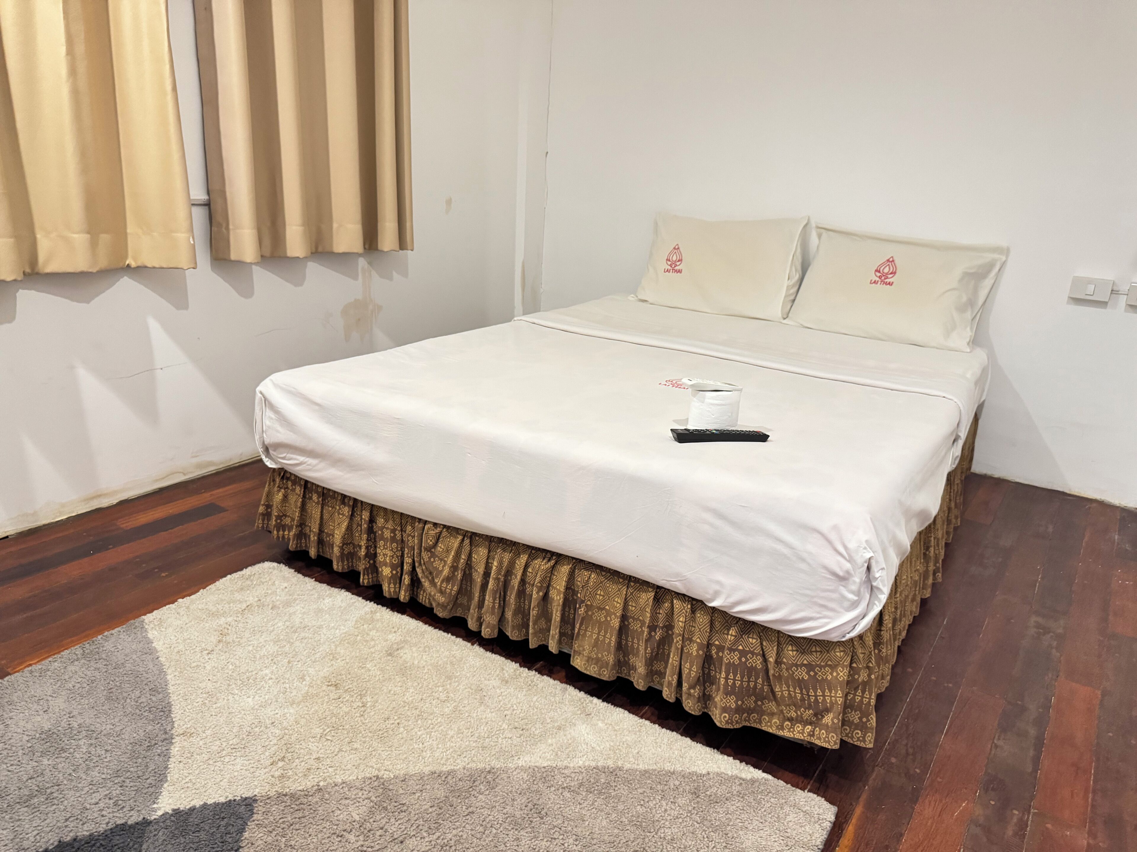 Una cama bien hecha con una colcha blanca y una falda estampada, un control remoto y una toalla enrollada sobre la cama.
