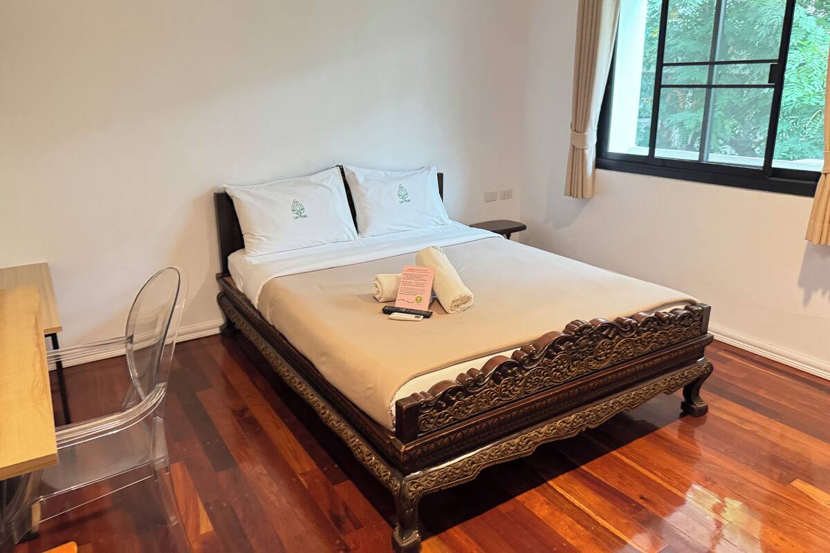Een net opgemaakt bed met een beige sprei en twee kussens, een houten nachtkastje met een lamp, en een raam met gordijnen.