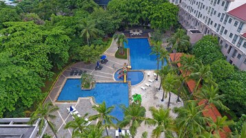 Uma piscina grande ao ar livre rodeada por palmeiras e espreguiçadeiras, com uma área de recreação infantil e um edifício ao fundo.