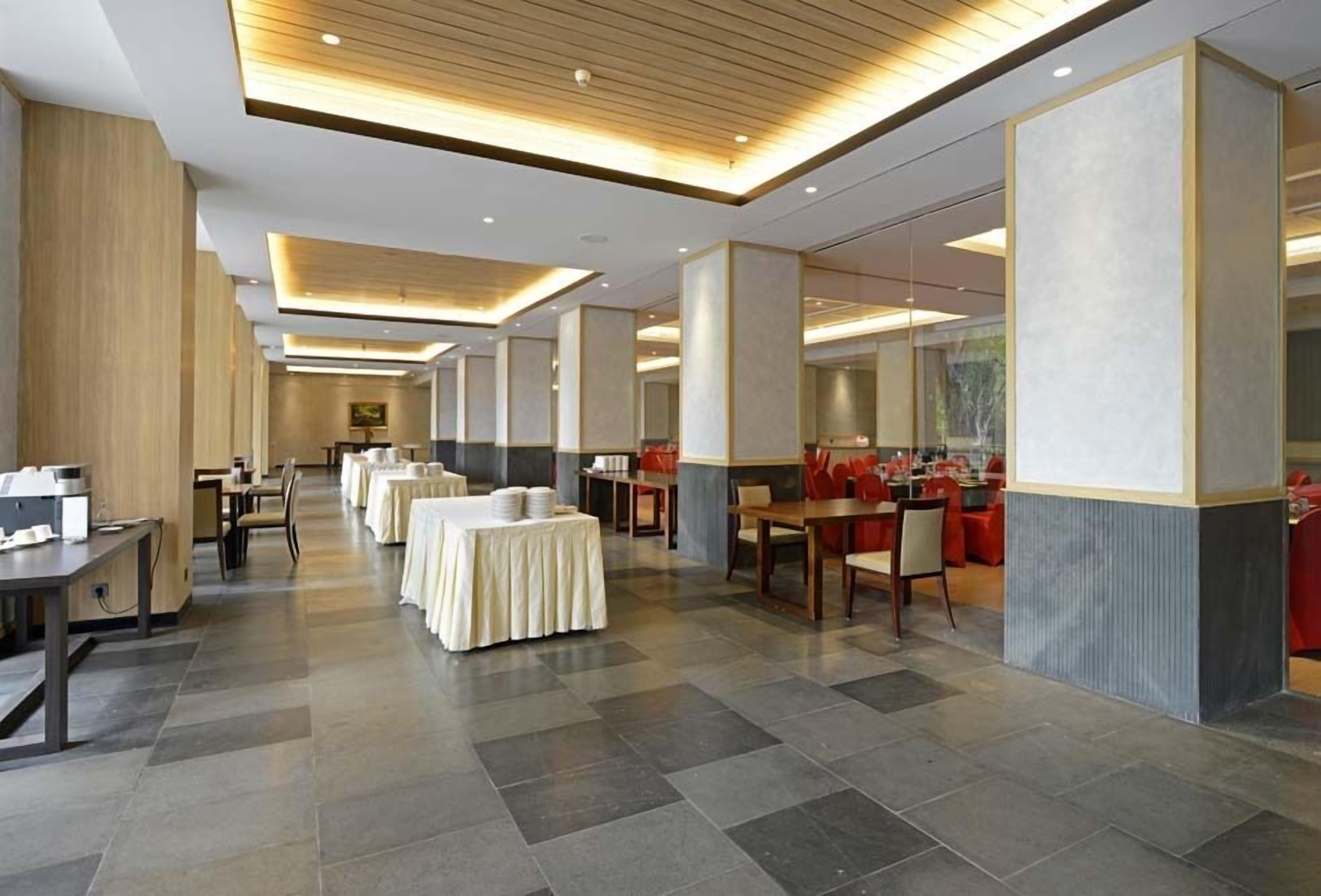 A spacious hotel lobby with multiple dining areas, including a buffet setup and several tables with chairs.