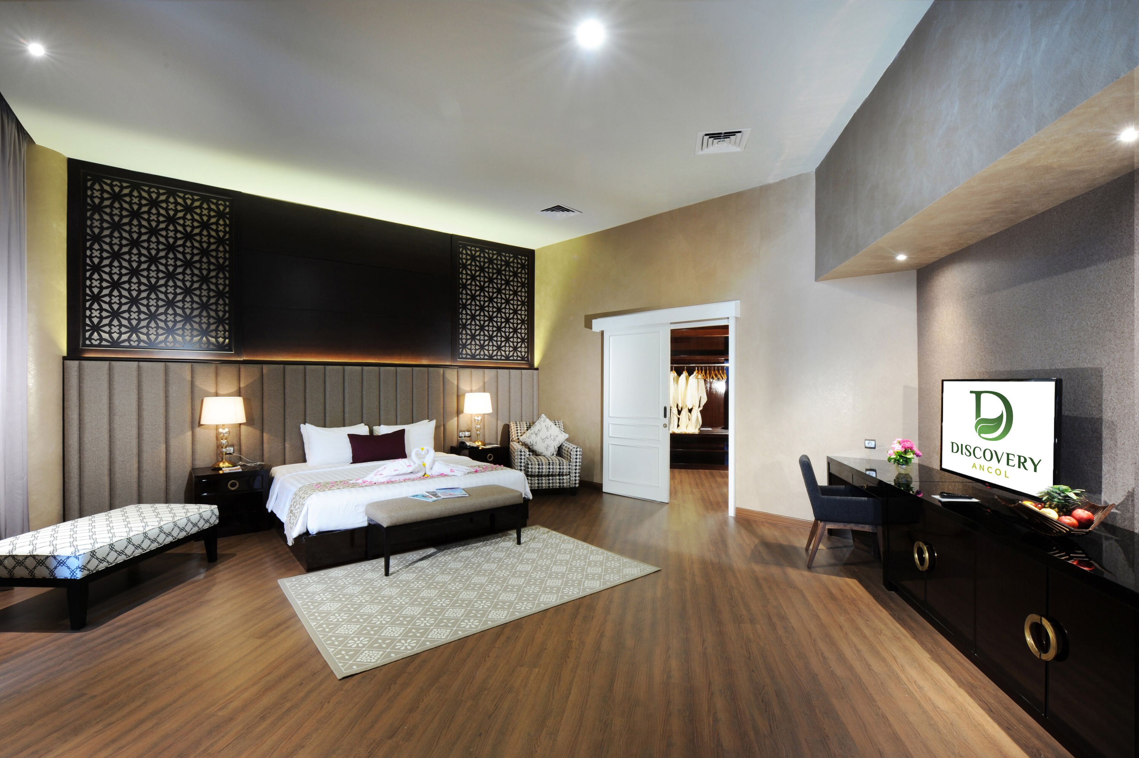 Quarto de hotel moderno com uma cama grande, mesas de cabeceira com luminárias, uma escrivaninha com um computador e um biombo decorativo.