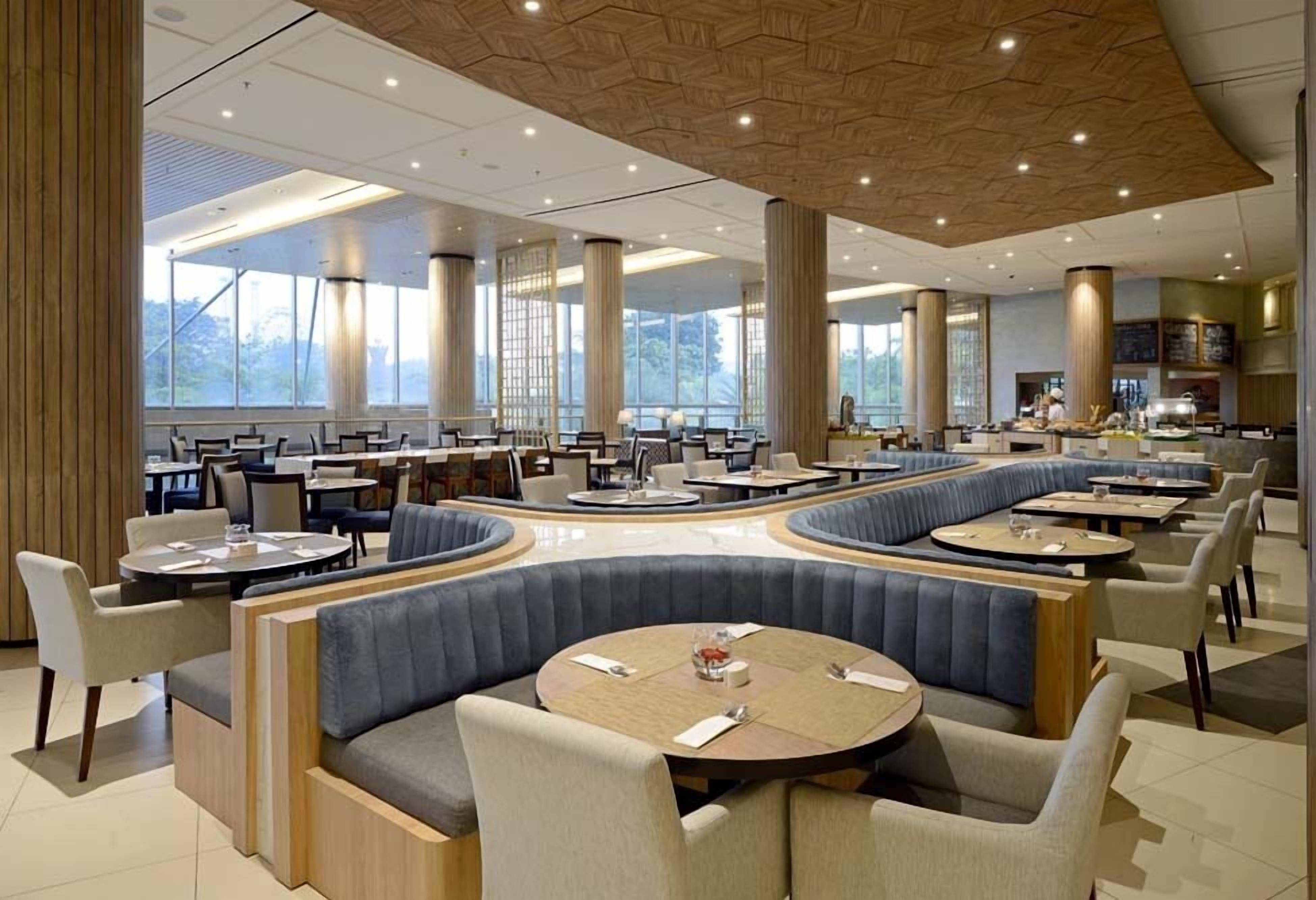 Un restaurante moderno con mesas redondas, asientos tapizados en azul y ventanas amplias que ofrecen vistas al exterior.