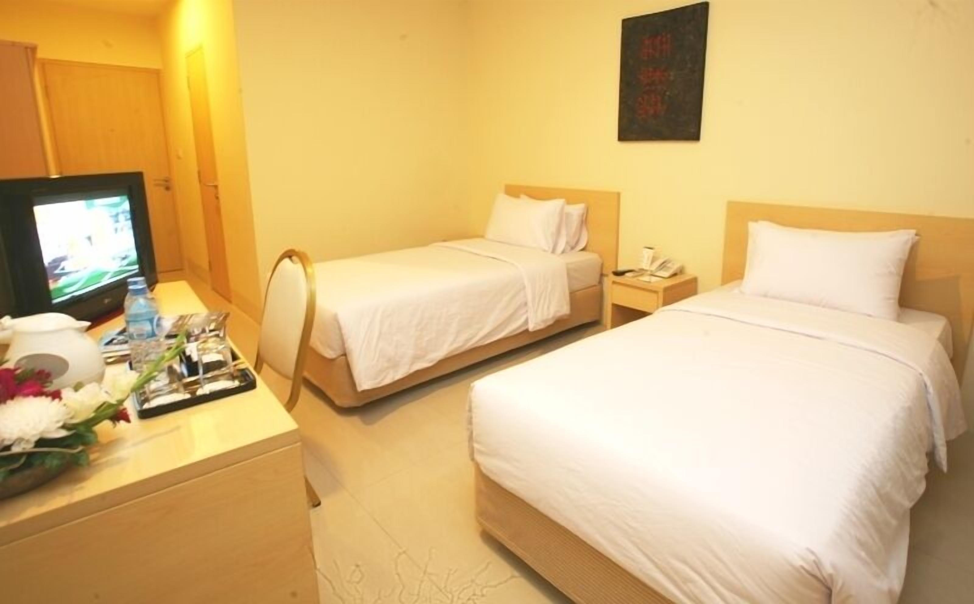 A hotel room with two single beds, a desk with a television, and a small table with a vase of flowers.