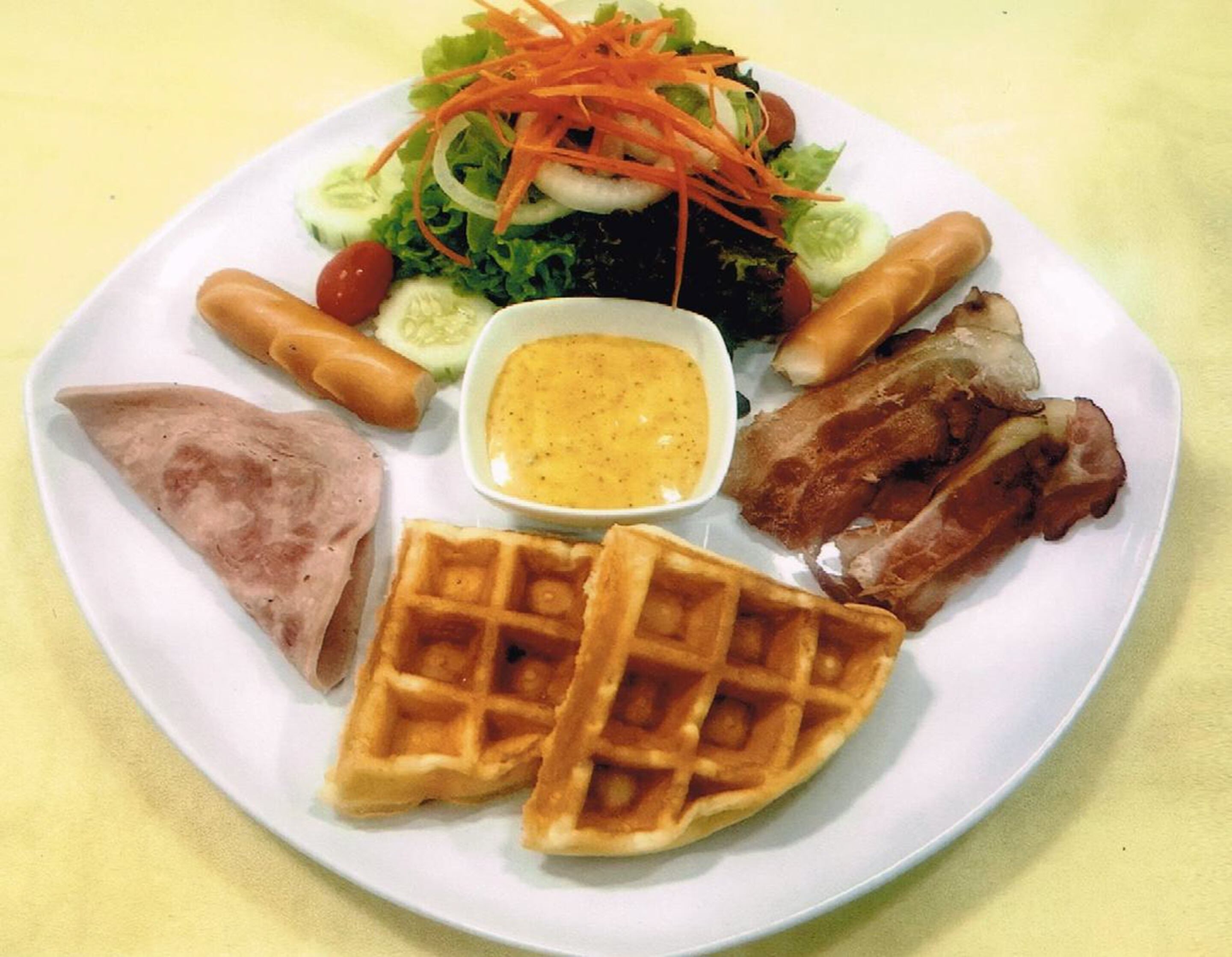 A plate with a variety of foods including a salad, sausages, a piece of meat, a waffle, and a small bowl of sauce.