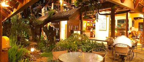 An indoor garden with a round table and wicker chairs, surrounded by tropical plants and a wooden structure.
