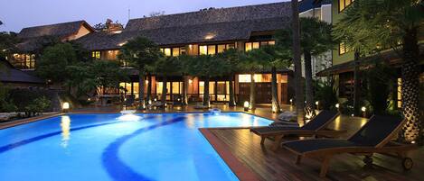 A swimming pool with lounge chairs, surrounded by buildings with thatched roofs and illuminated windows.