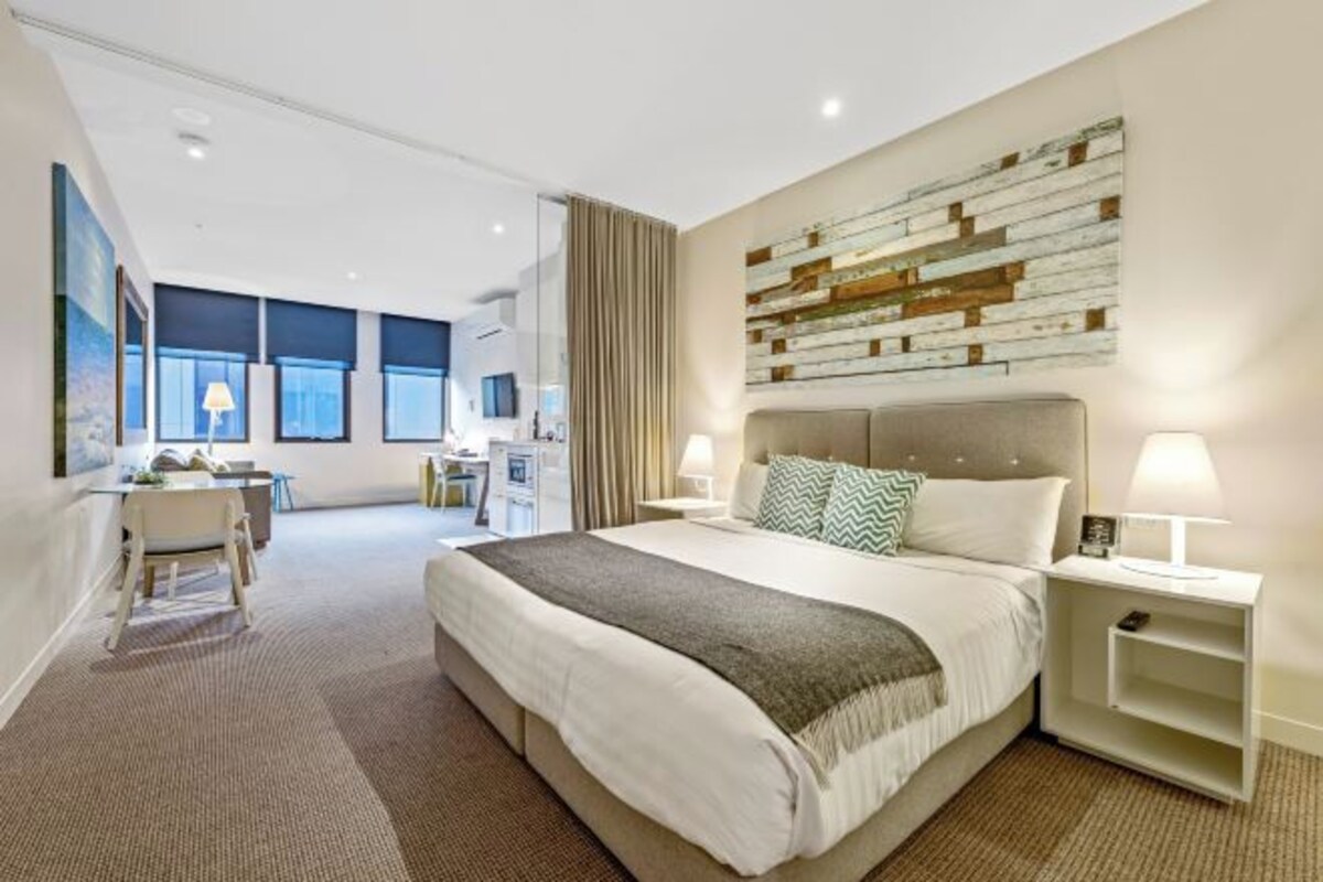 Habitación de hotel moderna con una cama grande, un escritorio y una silla. Se ve la ciudad a través de amplias ventanas.