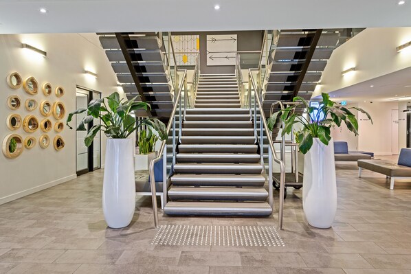 Un moderno vestíbulo de hotel con una escalera central, plantas en macetas y señalización.