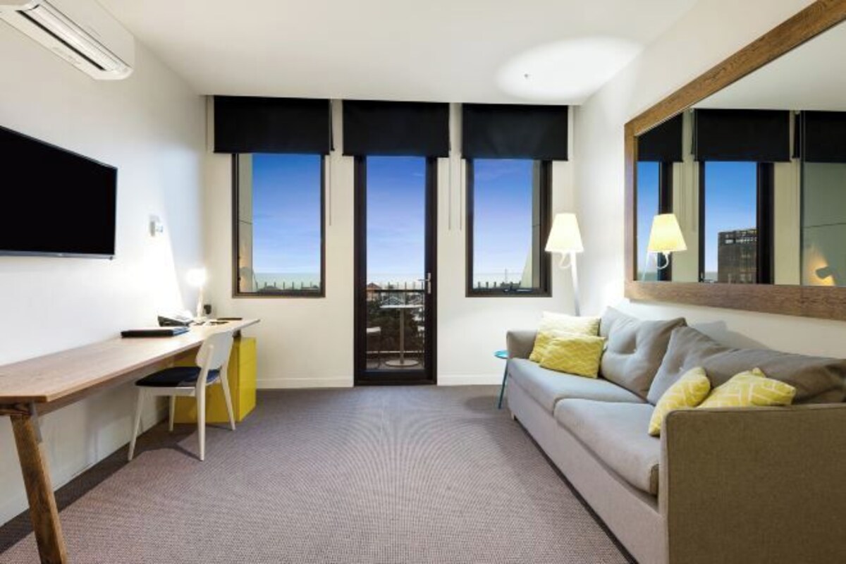 Habitación de hotel con escritorio, sofá, espejo y vistas de la ciudad a través de amplias ventanas.
