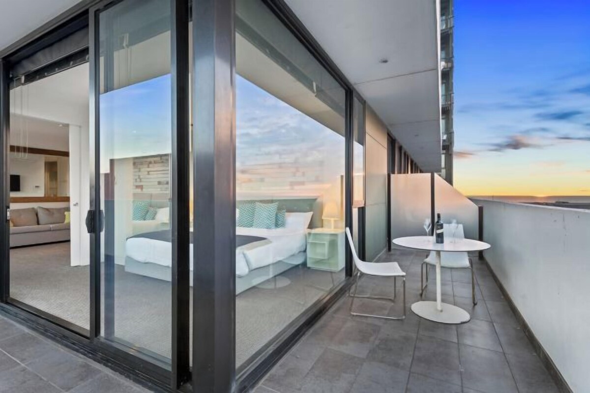 A modern balcony with a glass door leading to a bedroom, a round table, and a chair.