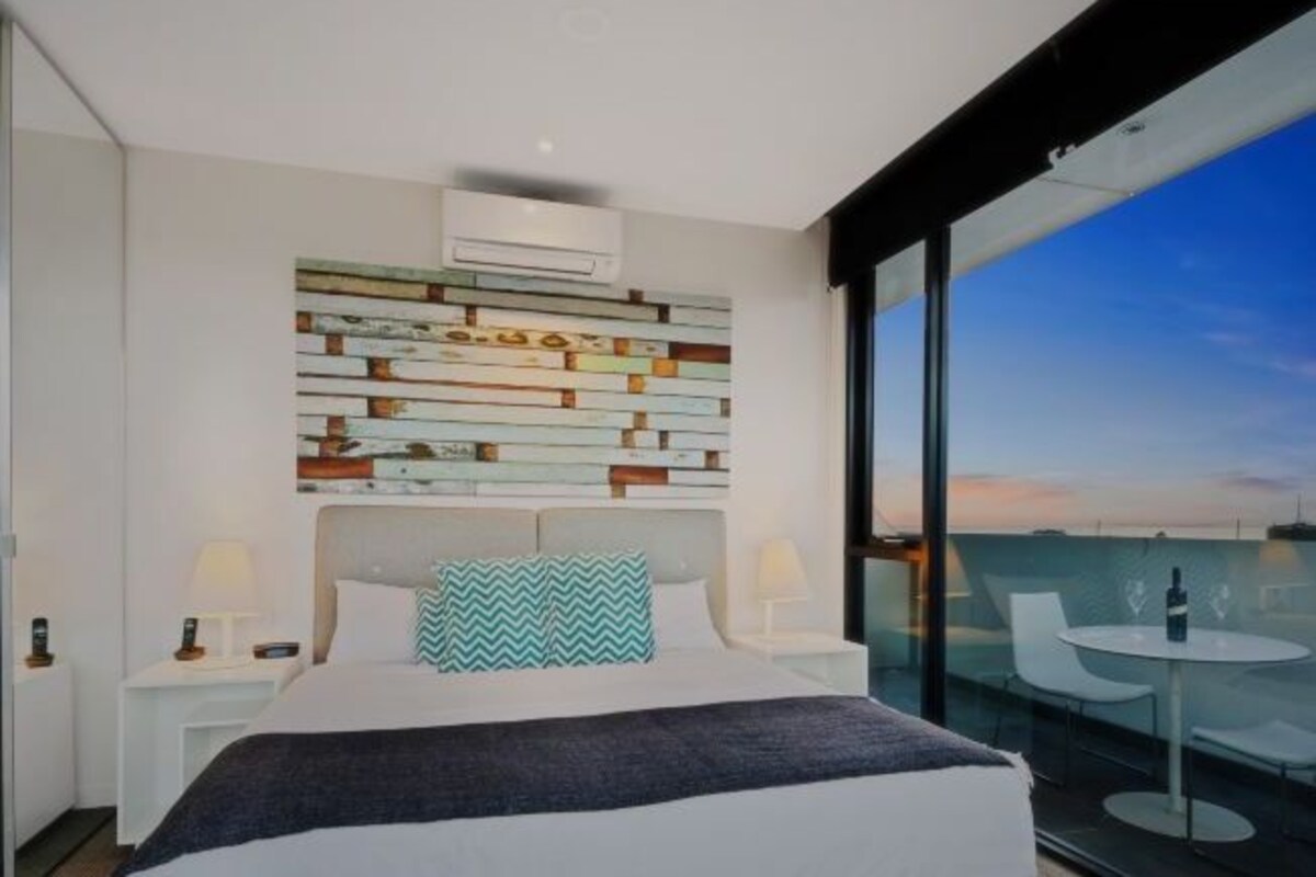 Un moderno cuarto de hotel con una cama grande, una pared decorativa, un balcón con una mesa y sillas de vidrio, y vista al horizonte de la ciudad.