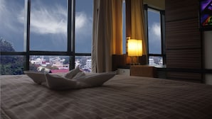A hotel room with a large bed, a nightstand, a lamp, and a view of the city through large windows. - Travello Manado (Manado)
