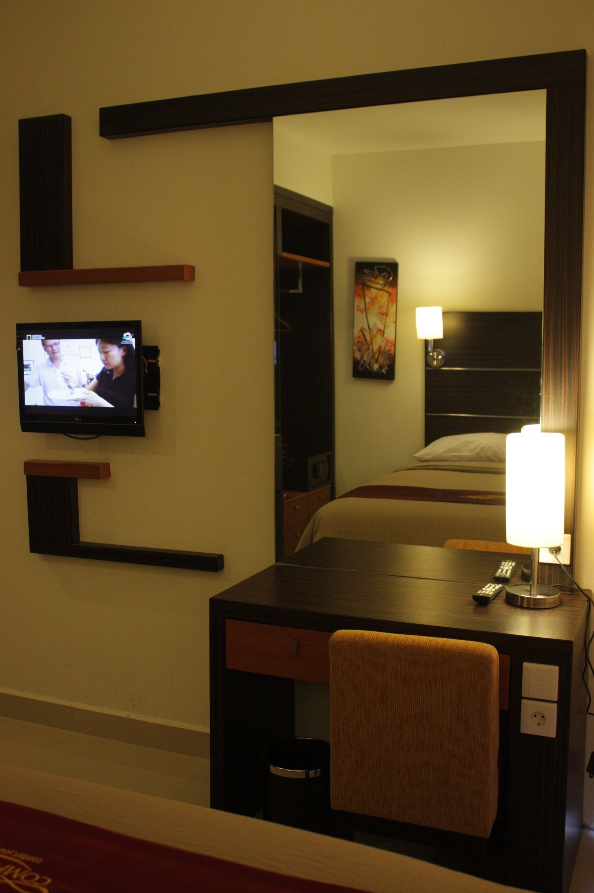 A hotel room with a television mounted on the wall, a desk with a chair, a bedside lamp, and a bed with a headboard.