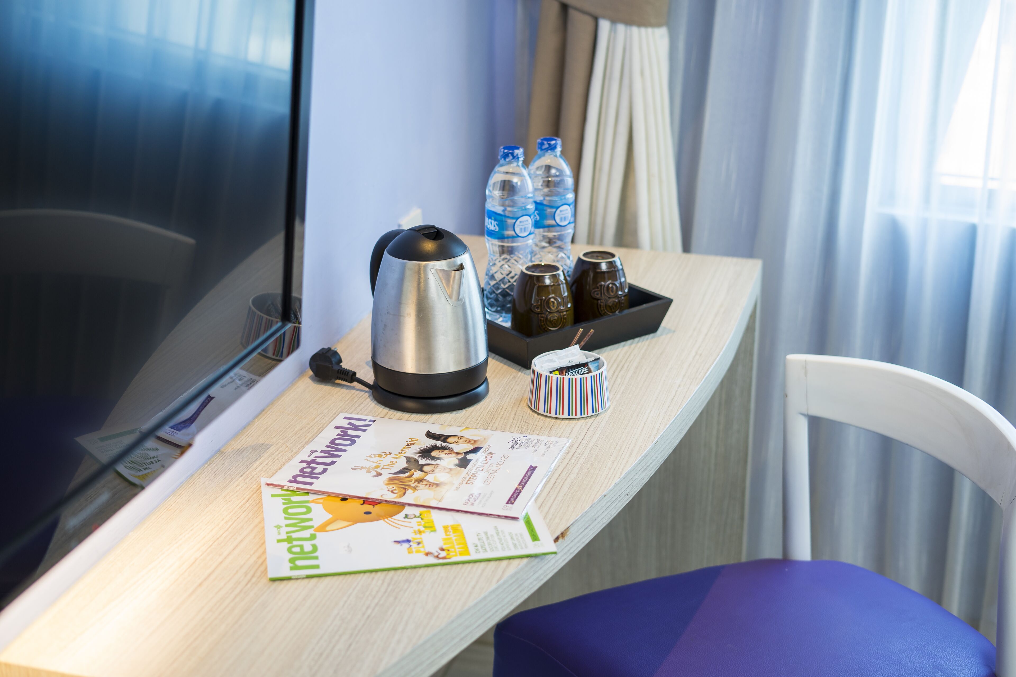 A hotel room with a wooden desk, a kettle, water bottles, salt and pepper shakers, a magazine, and a purple chair.