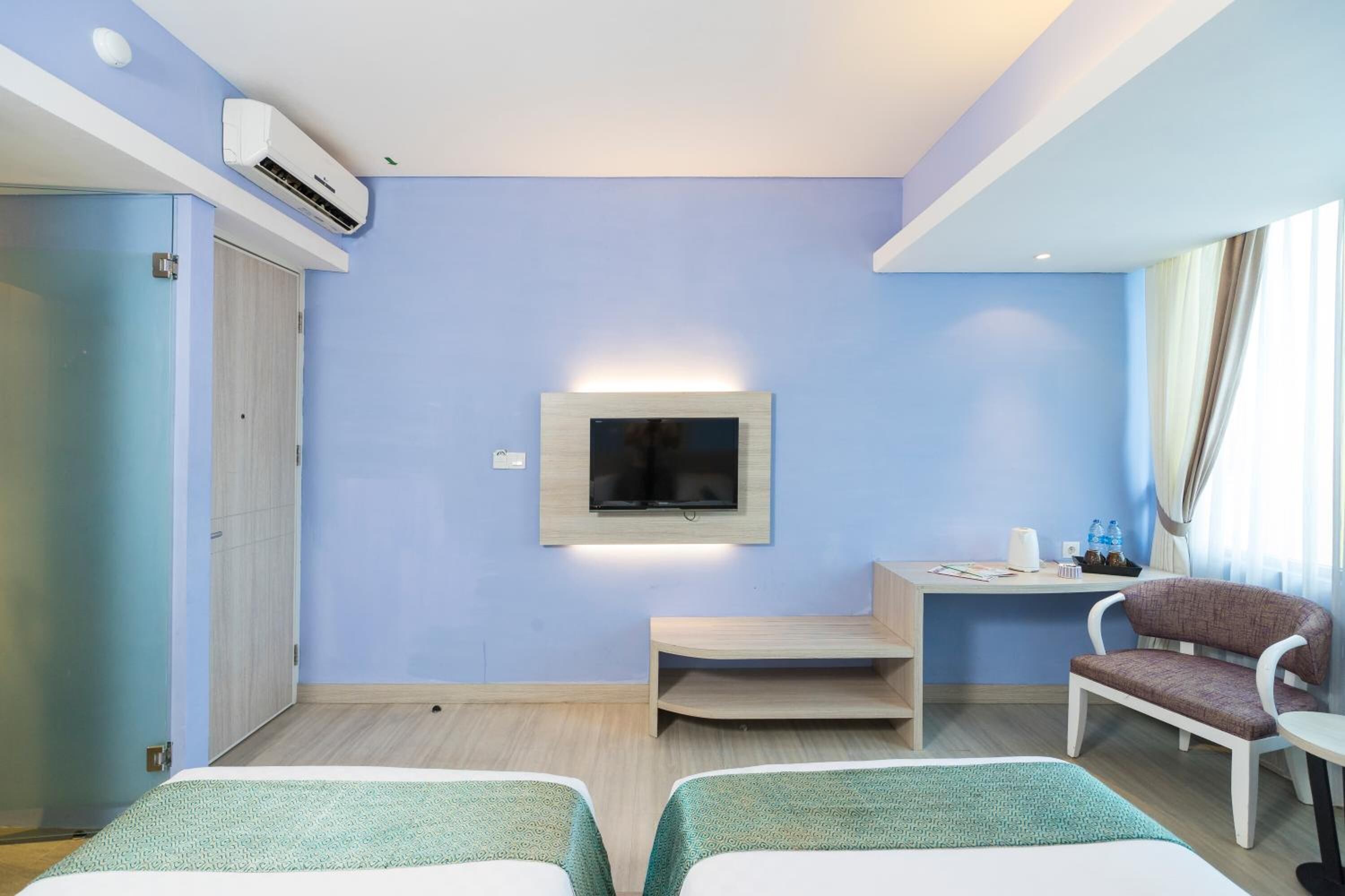 Une chambre d’hôtel avec deux lits, un téléviseur fixé au mur, un bureau avec une chaise et une fenêtre avec des rideaux.