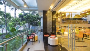Un espace intérieur moderne, aménagé avec des coins salon, une balustrade en verre donnant sur un espace verdoyant, et un espace café avec des tables et des chaises.