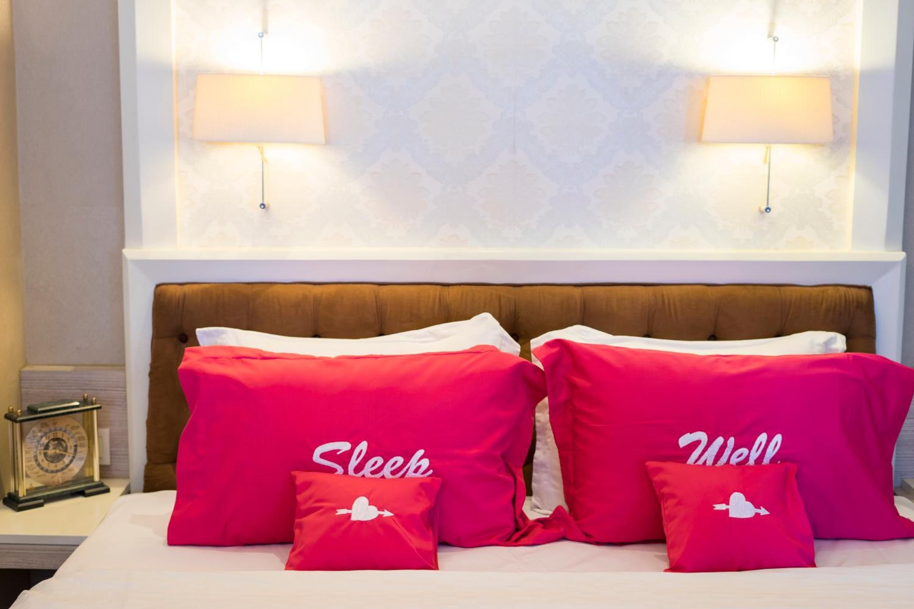 A hotel room with a bed, red pillows with white text, two wall-mounted lamps, and a clock on the nightstand.
