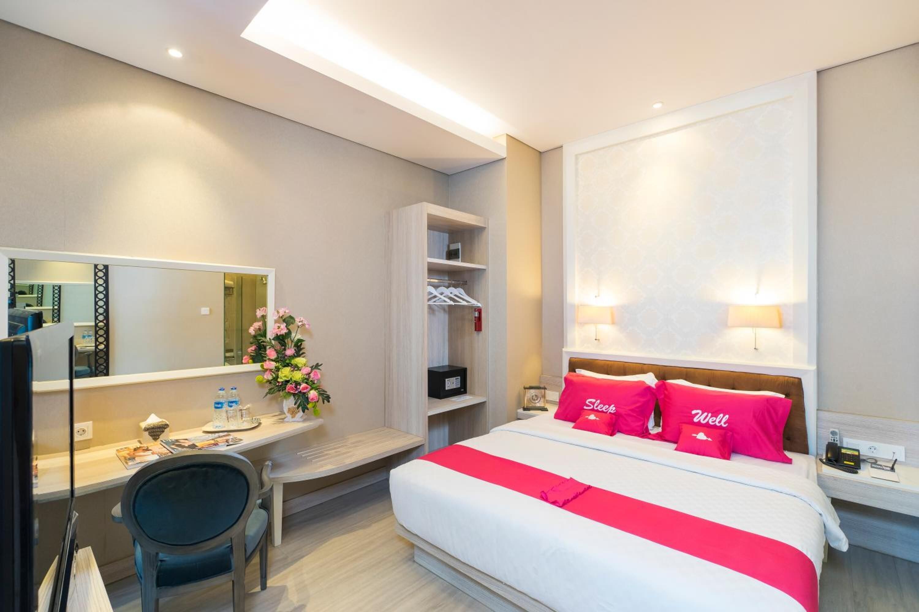 A hotel room with a bed, bedside lamps, a desk with a chair, a mirror, a shelf with toiletries, and a vase with flowers.