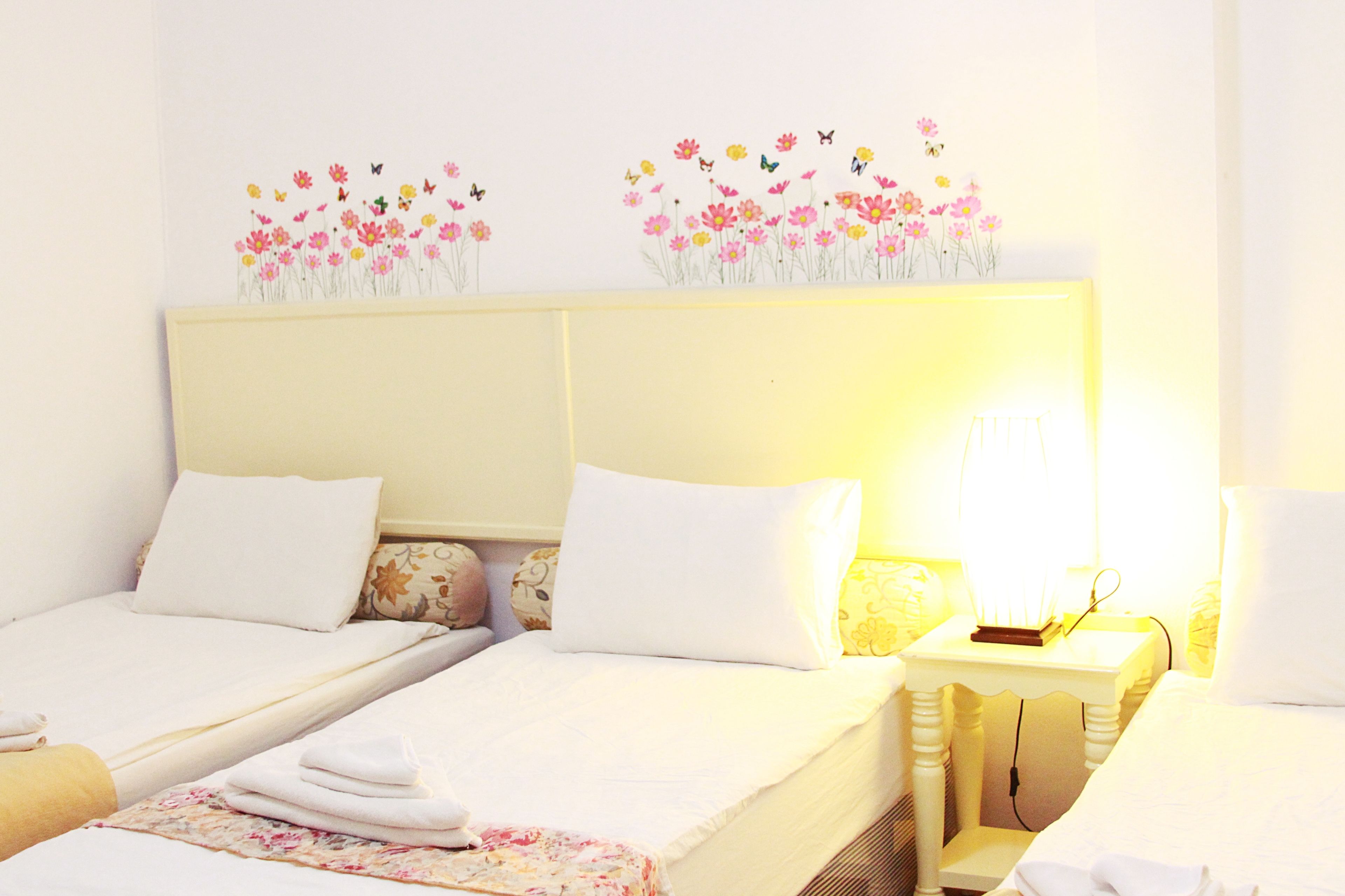 A hotel room with two beds, each with white linens and floral-patterned pillows. There is a bedside table with a lamp and a book on one side.