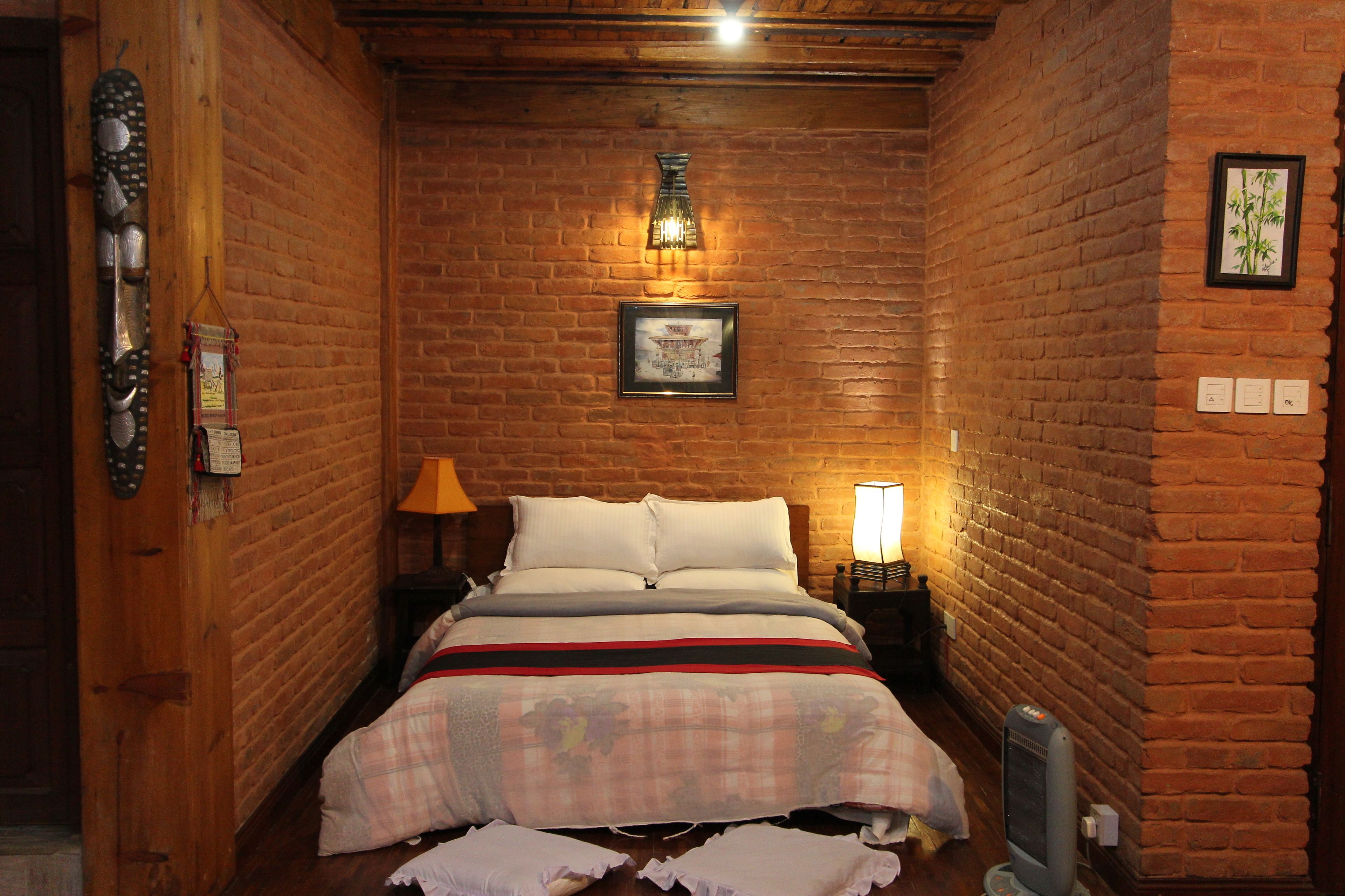 A narrow room with a brick wall, a bed with a plaid bedspread, a wall-mounted lamp, and a framed picture.