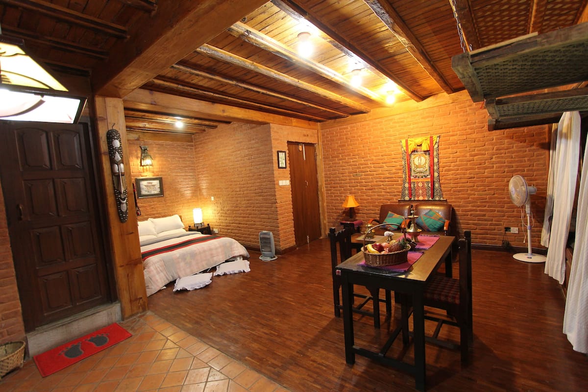 Een kamer met een houten plafond, bakstenen muren, een bed met een witte dekbedovertrek, een tafel met stoelen en een ventilator.