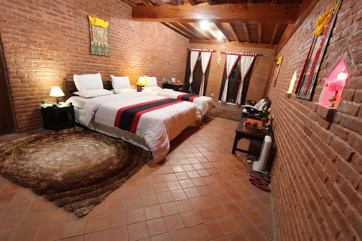 Een kamer met een bakstenen muur, twee bedden met witte beddengoed en rode accenten, een bruin vloerkleed en een klein tafeltje met een lamp.