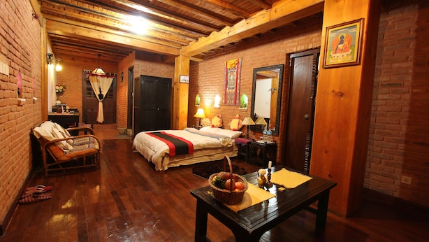 A cozy room with a wooden ceiling, a bed with a red and black bedspread, a wicker chair, and a table with a basket of fruit. - World Heritage Hotel & Aparments (Kathmandu)