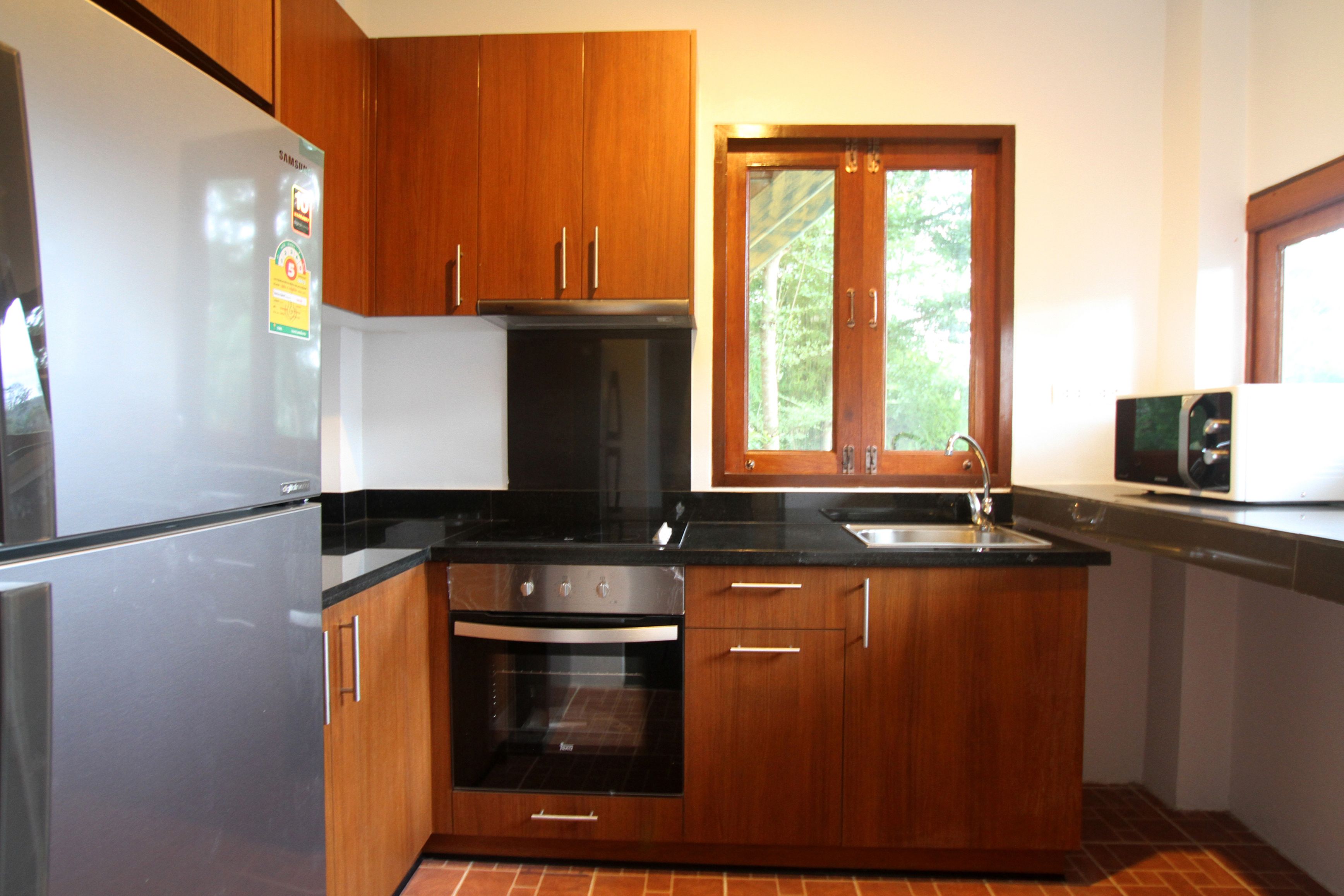 A kitchen with wooden cabinets, a stainless steel refrigerator, an oven, a sink, and a window with a view of trees.