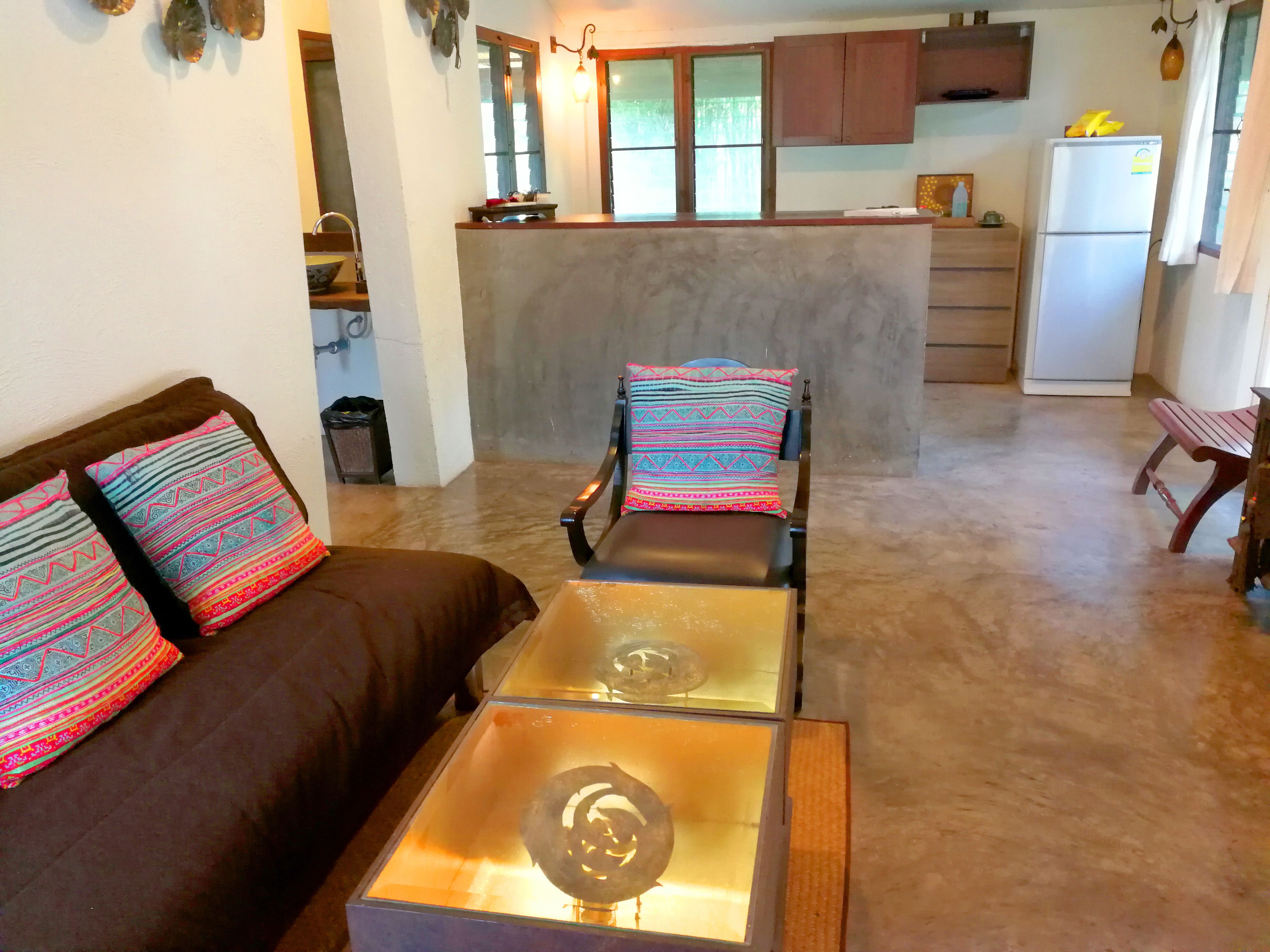 A living room with a sofa, a glass coffee table, and a chair with patterned cushions. There is a kitchen area in the background with cabinets and a refrigerator.
