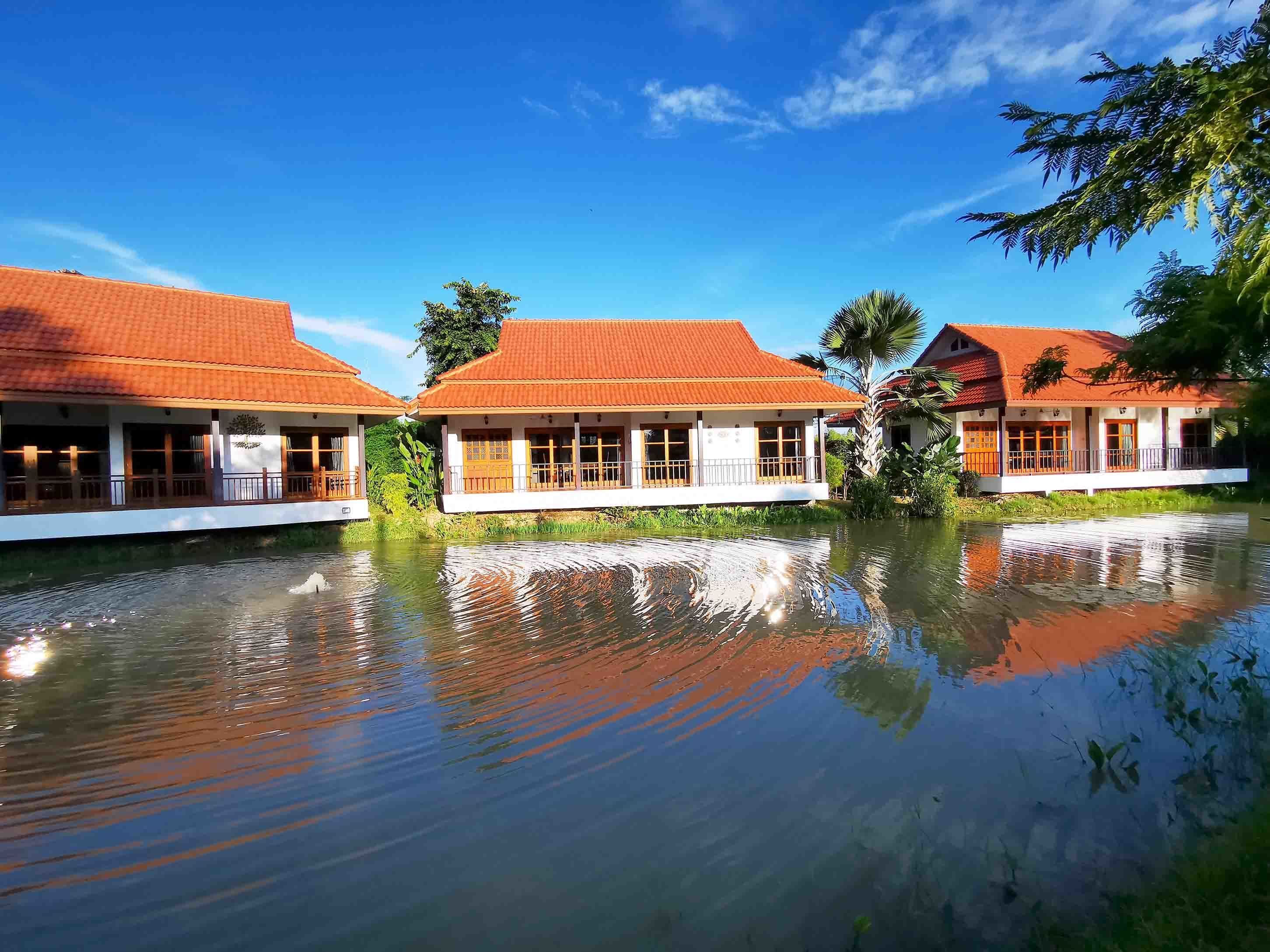 A row of traditional-style buildings with red roofs, white walls, and large windows, reflecting in a calm body of water.
