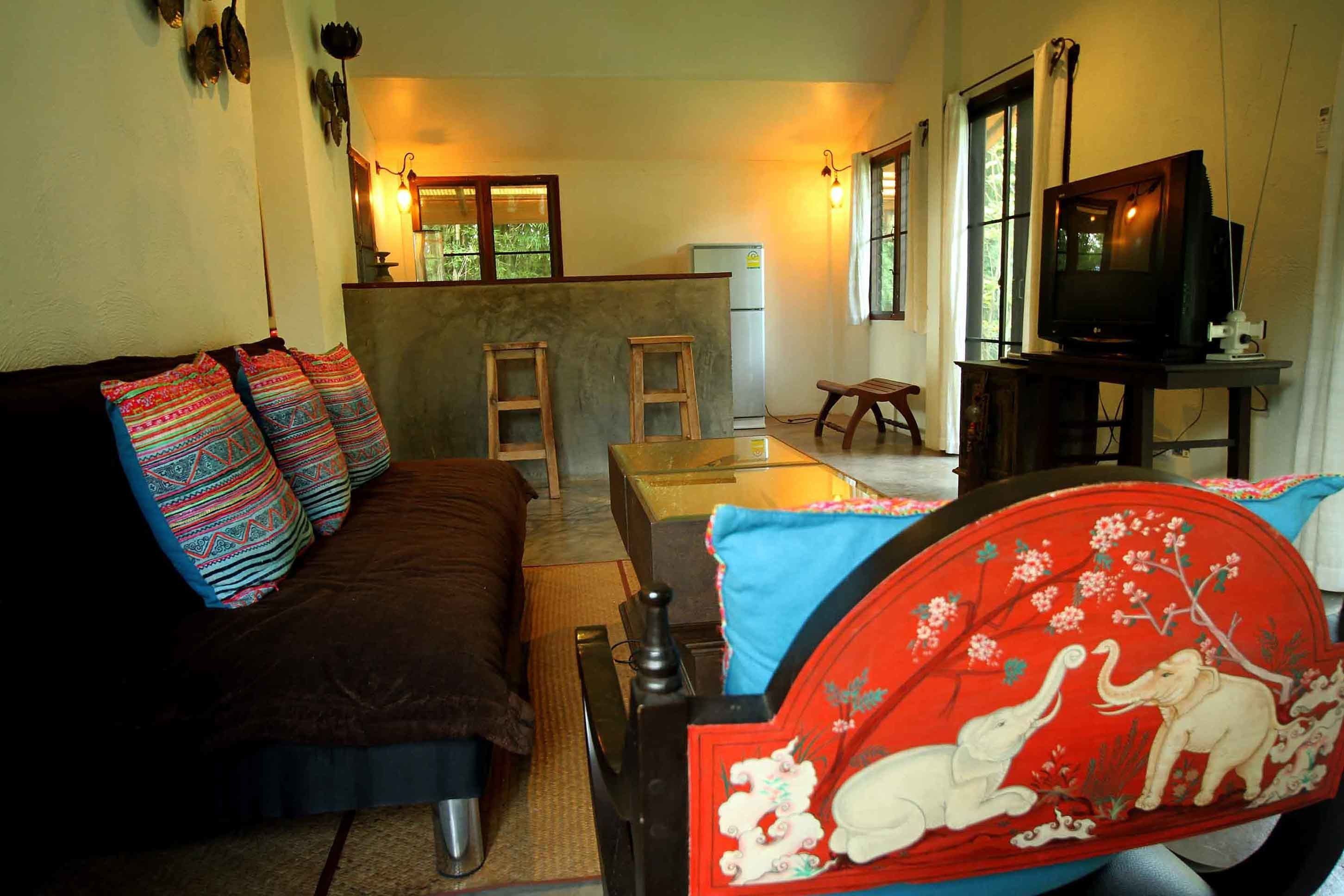 A cozy living room with a brown sofa, colorful pillows, a glass coffee table, a red and white elephant-themed headboard, a television, and a kitchen area in the background.