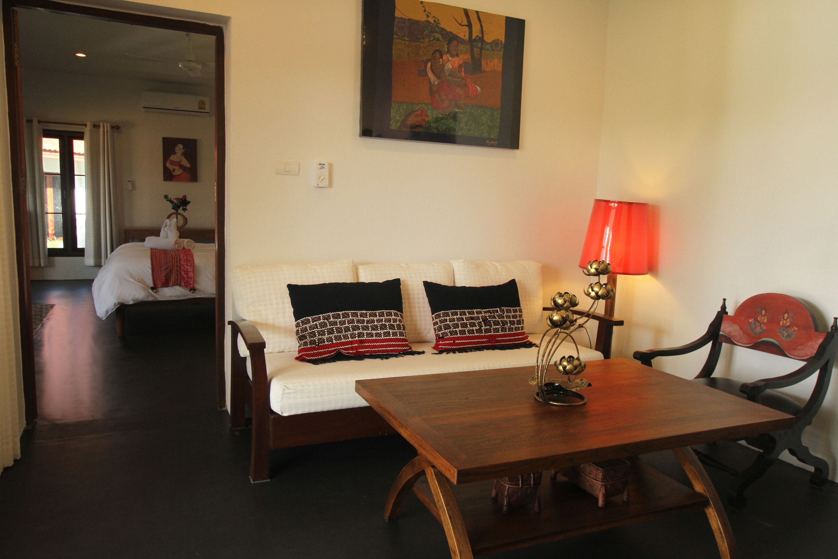 Una habitación con un sofá blanco, una mesa de madera y una lámpara roja. Hay un cuadro en la pared y una silla con un respaldo decorativo.