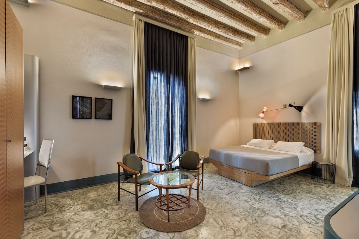 A hotel room with a bed, a round table with chairs, a small table with a bowl, and a wall-mounted lamp.