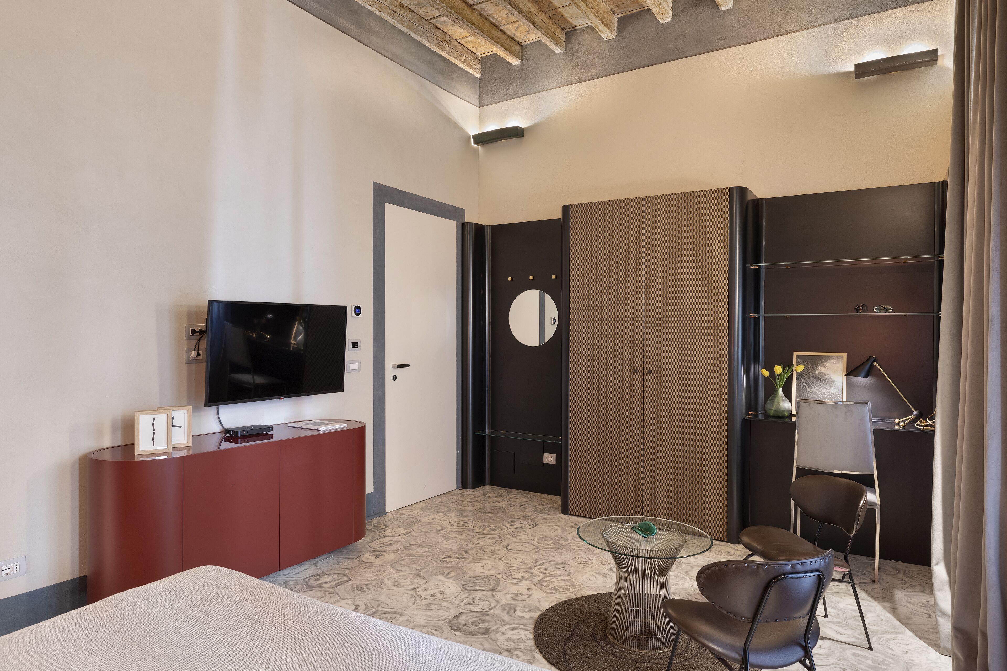 Una habitación de hotel moderna con televisión de pantalla plana, un minibar rojo, una mesa redonda con sillas, un espejo grande y una alfombra estampada.