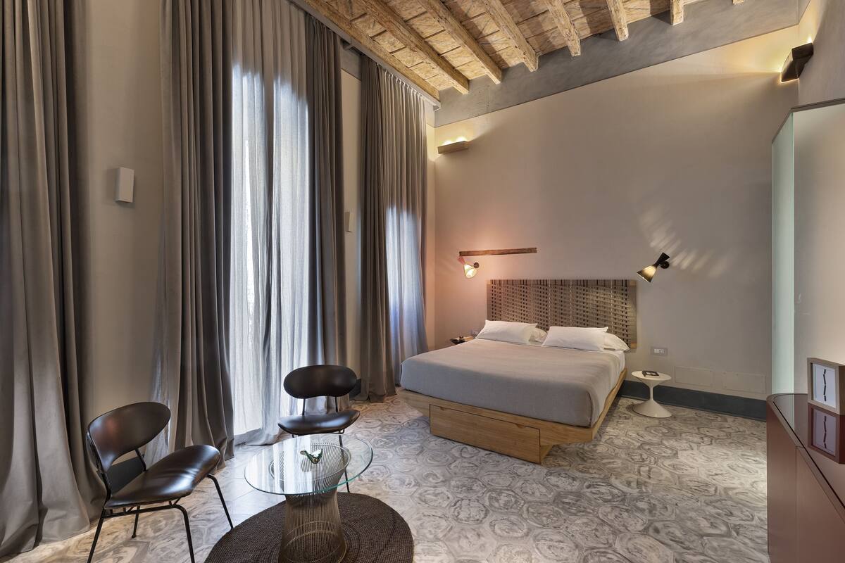 A modern hotel room with a large bed, a small round table, two chairs, a bedside table, and a large window with curtains.