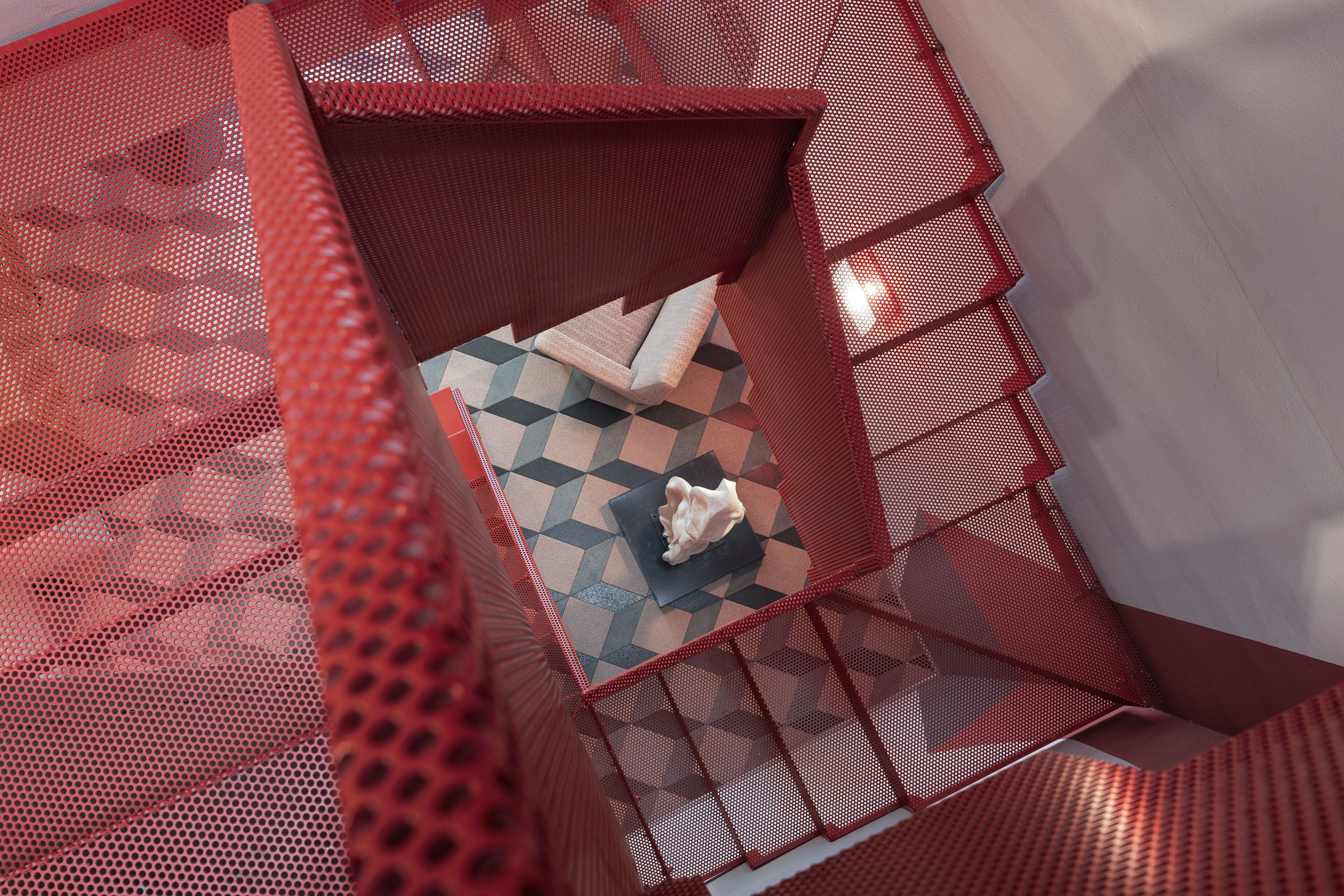 Un escalier en colimaçon avec des rampes en métal rouge et un sol orné d’un motif géométrique.