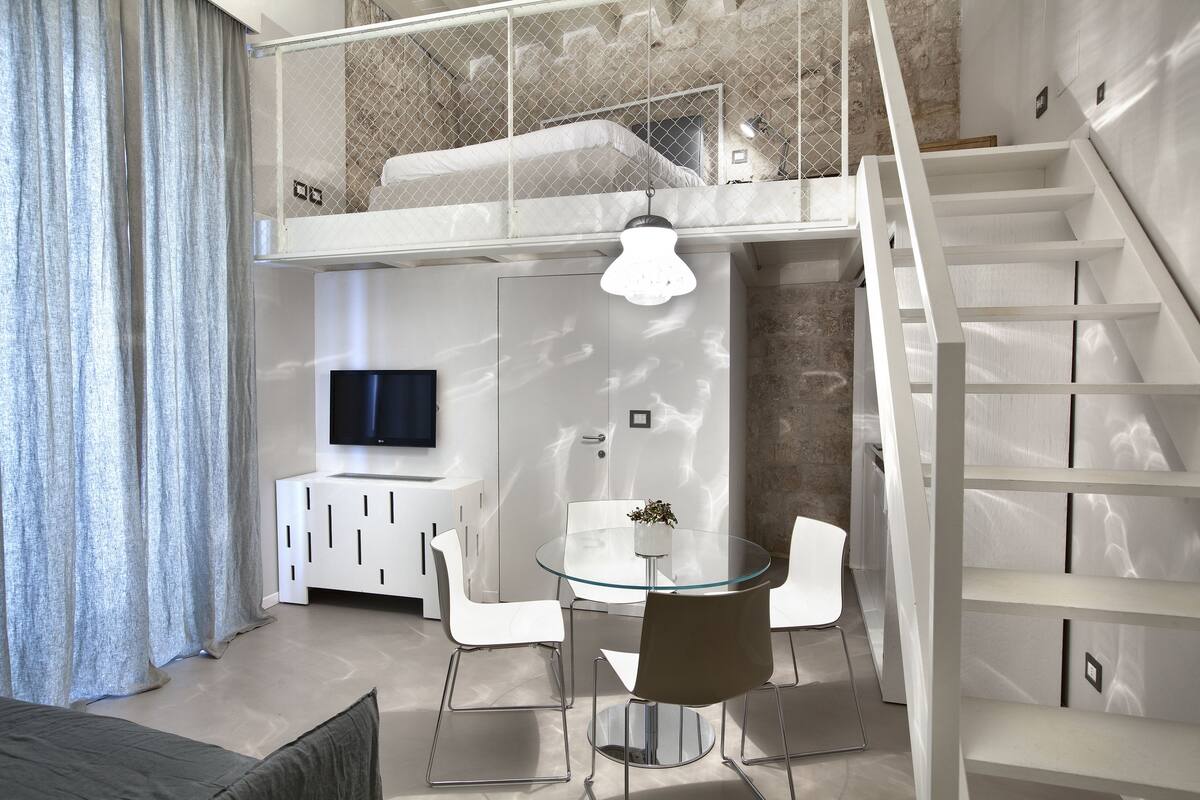 A modern living room with a glass dining table, white chairs, a TV, and a staircase.