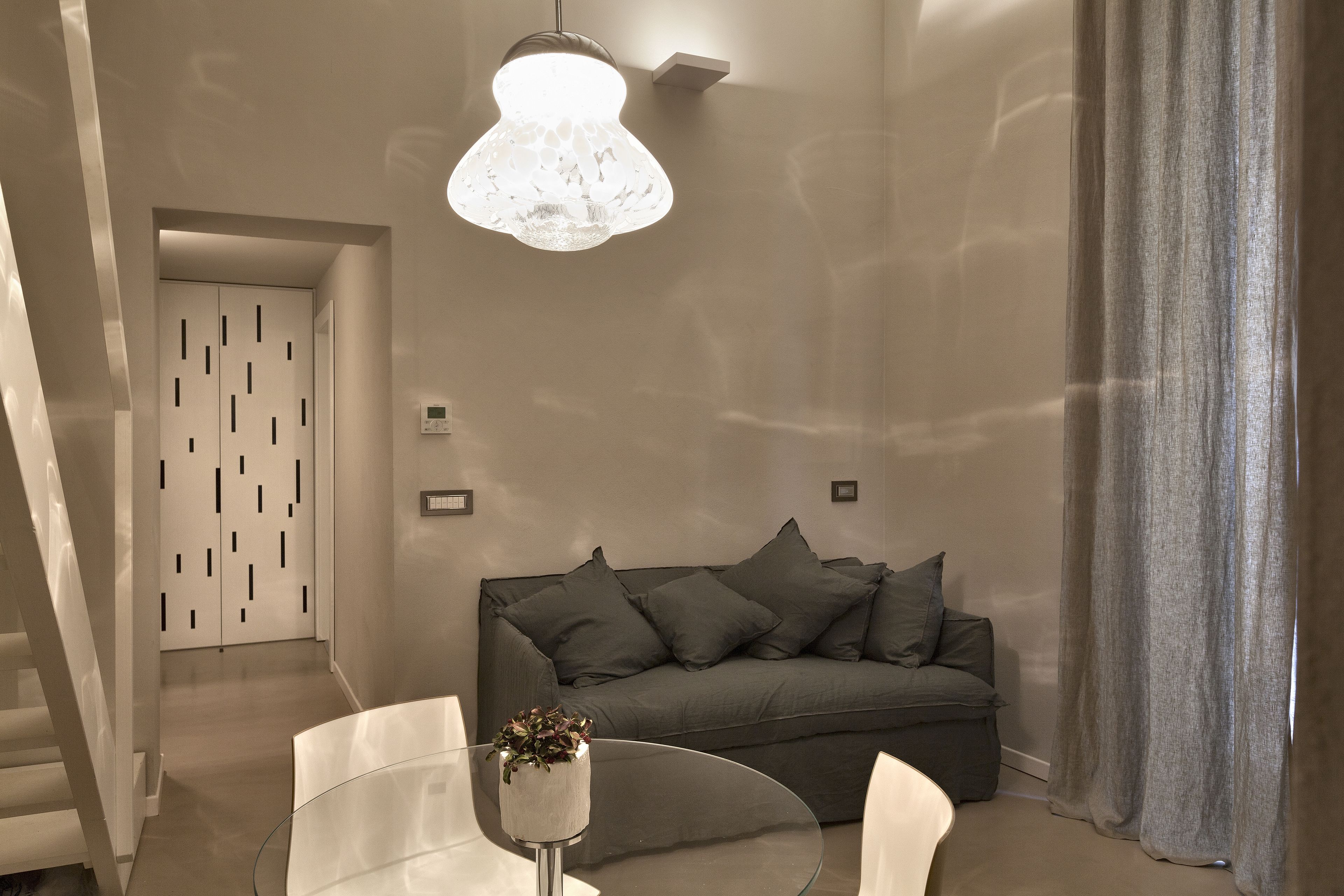 Una sala de estar moderna con una mesa de centro de vidrio, un sofá gris con varios cojines, una lámpara colgante y una puerta con un diseño geométrico.