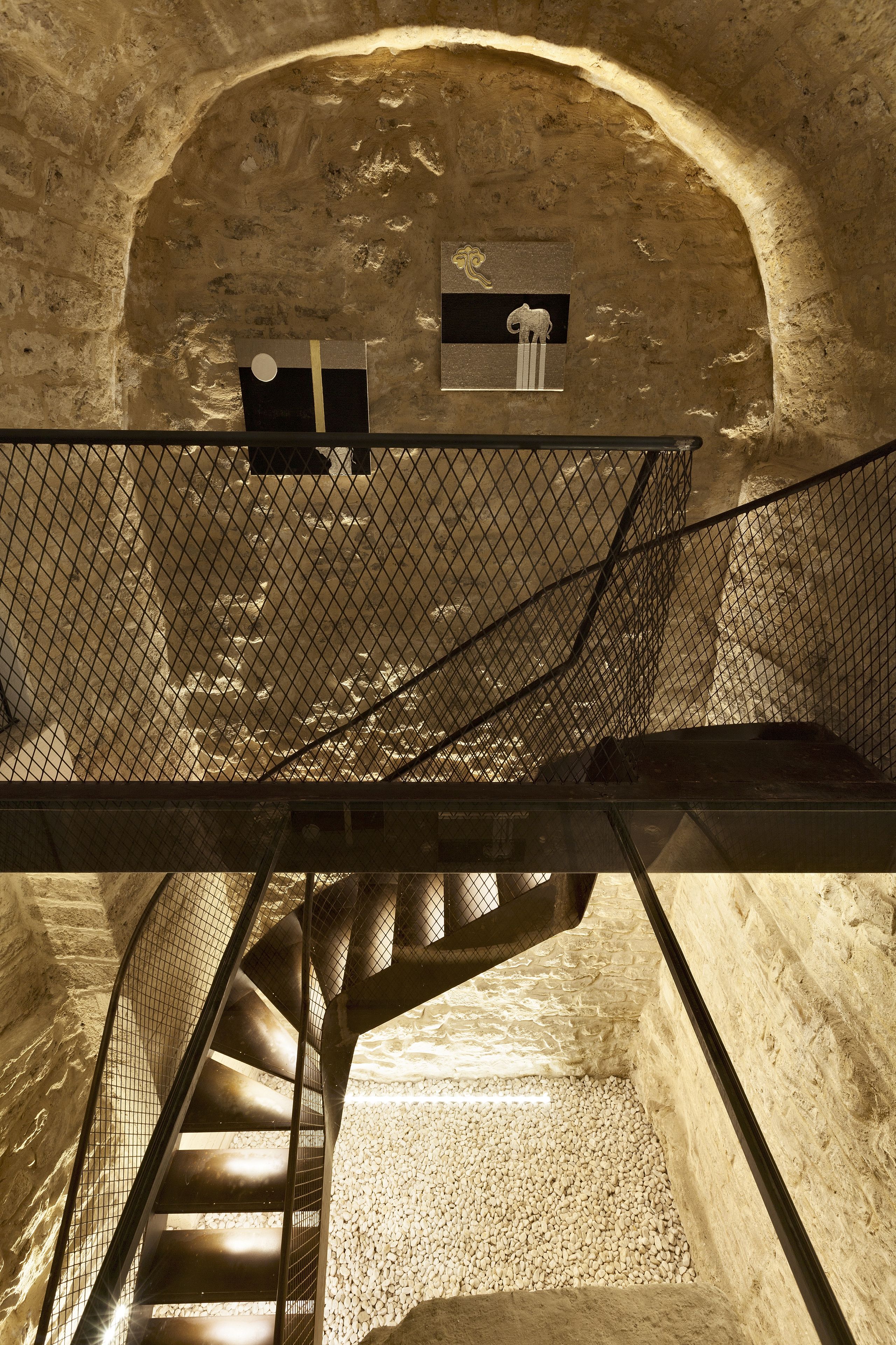 A spiral staircase in a stone-walled room with a metal railing and a small table with a chair.