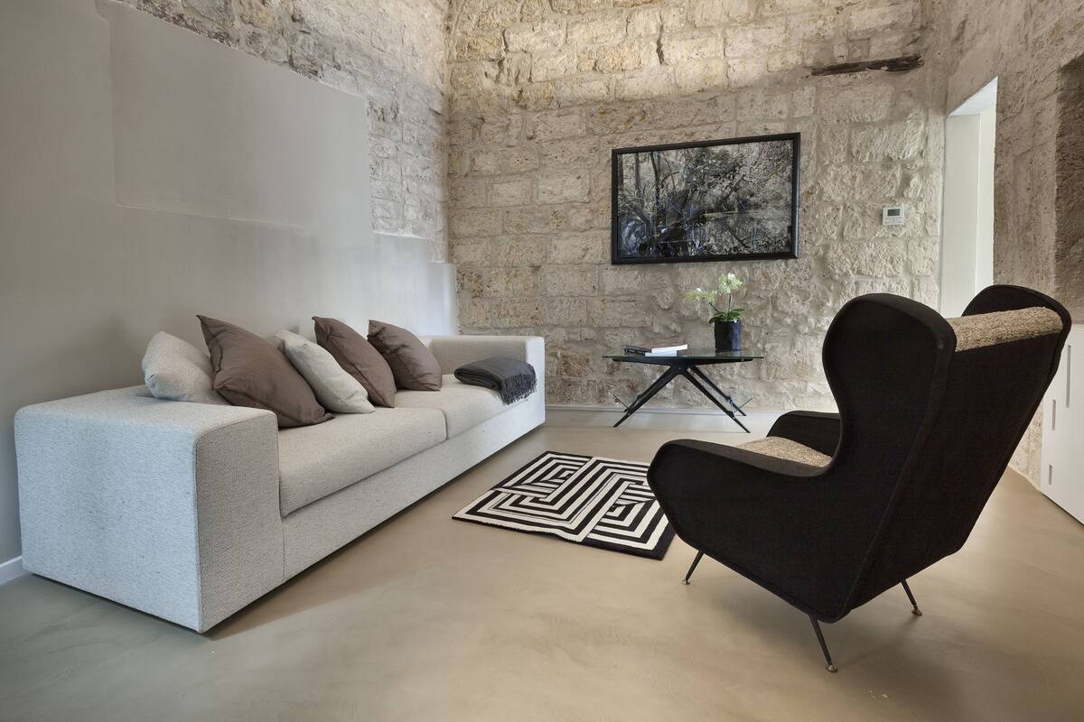 A modern living room with a stone wall, a white sofa with cushions, a black armchair, a glass coffee table, and a framed picture on the wall.