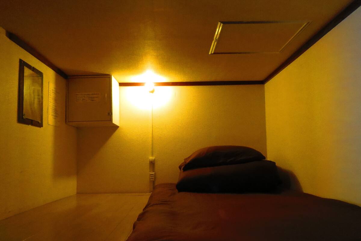 A small, dimly lit room with a single bed, a wall-mounted air conditioner, and a ceiling vent.